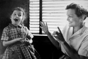 Little girl in a plaid jumper opens her mouth wide to speak next to a squatting woman smiling at her with her hands raised in communication with her.