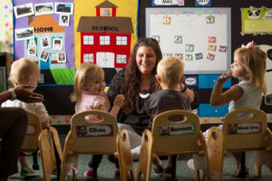 Four very young children seen from the back and facing forward to see their teacher's face in a bright classroom at Magnolia Speech School.