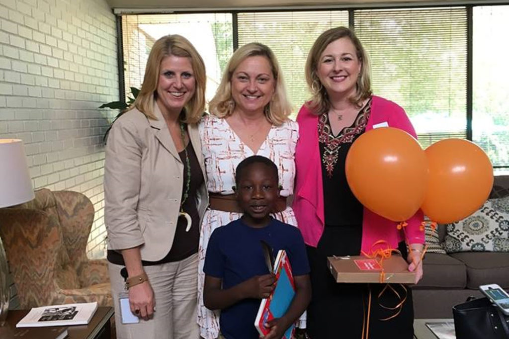 Three women stand and smile arm in arm. The woman on the right holds two orange balloons. A little boy stands in front, smiling at the camera.