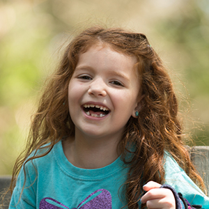Young girl with curly red hair wearing a teal shirt smiles into the camera.