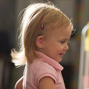 Blonde, smiling girl toddler in profile wearing a pink polo shirt and a hearing device.